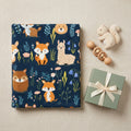A professional flat-lay gift presentation of the navy blue woodland baby blanket, folded neatly next to a wooden baby rattle, a small plush squirrel, and an elegant gift box. Perfect baby shower gift setup, soft warm studio lighting.