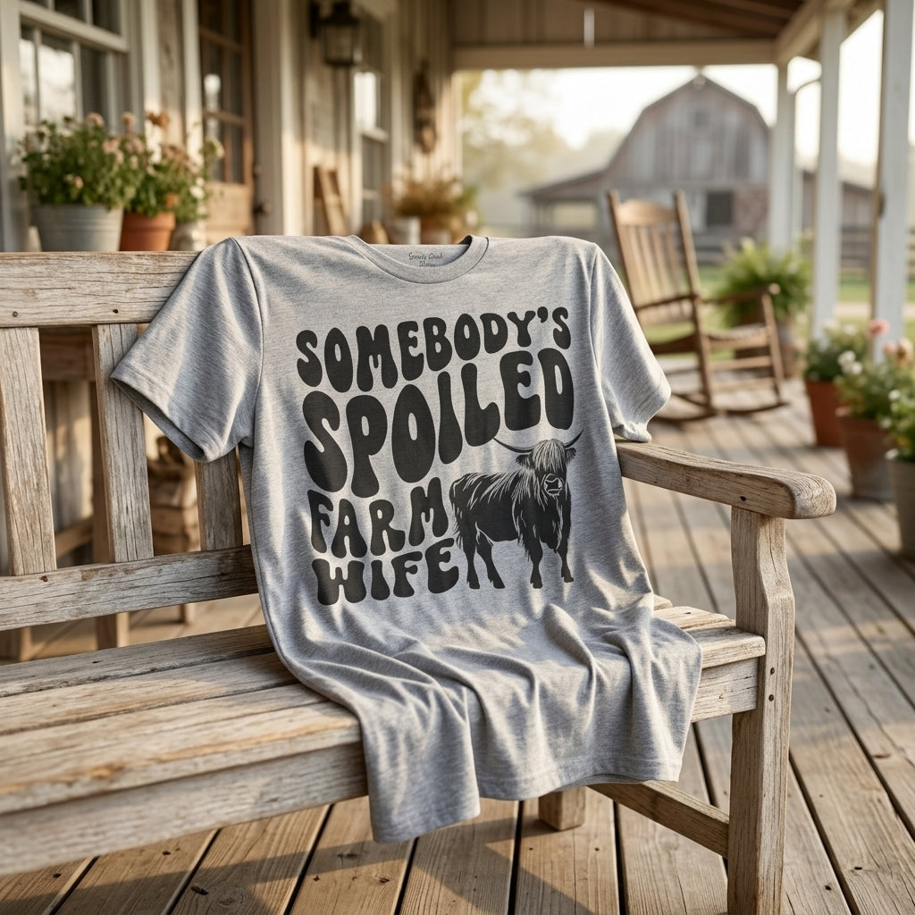 A rustic lifestyle scene showing the heather gray Highland Cow Spoiled Farm Wife t-shirt draped on a porch bench in warm morning sunlight