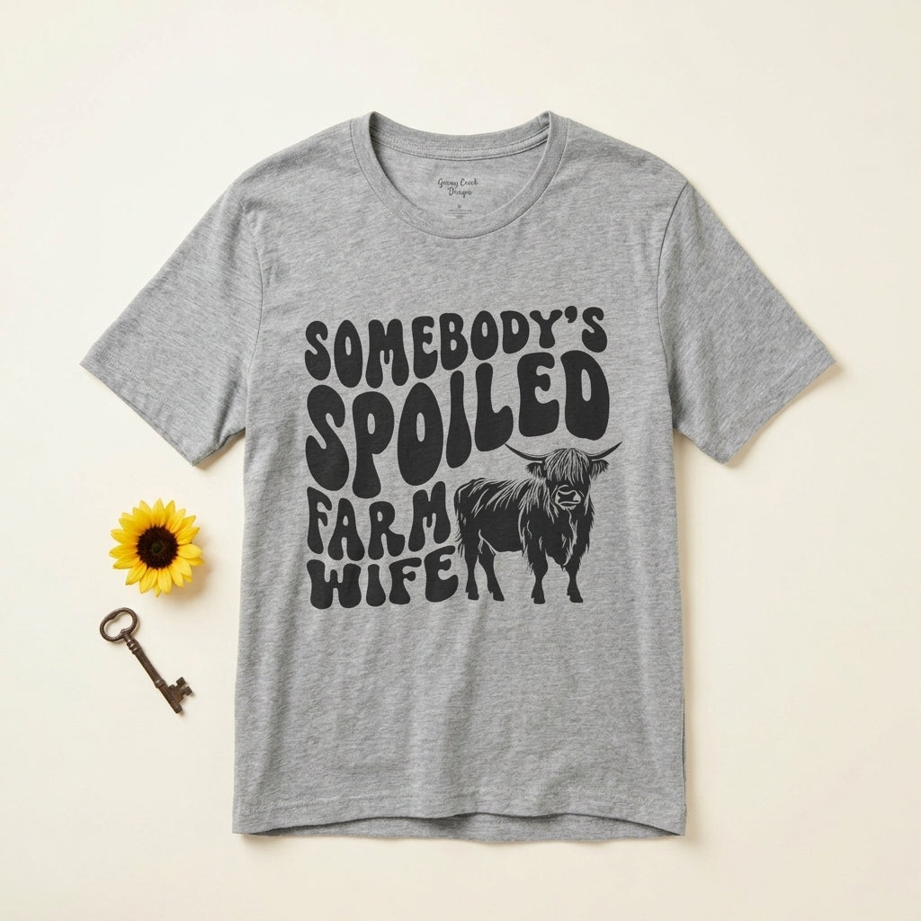 An e-commerce infographic-style image featuring the Highland Cow Spoiled Farm Wife t-shirt on a warm cream-colored background with props